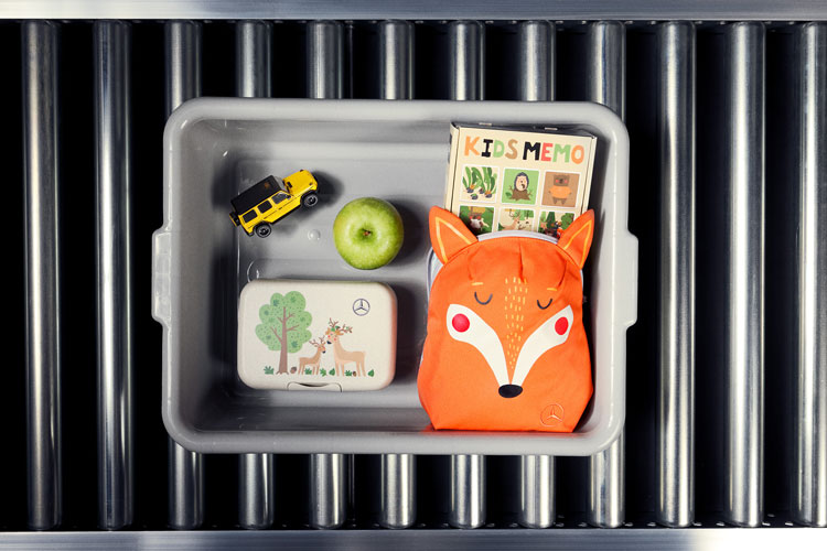 Mercedes-Benz Kids Collection Abgebildet ist eine graue Plastikbox auf einem Rollband, in welcher neue Artikel der Mercedes-Benz Kinder Kollektion